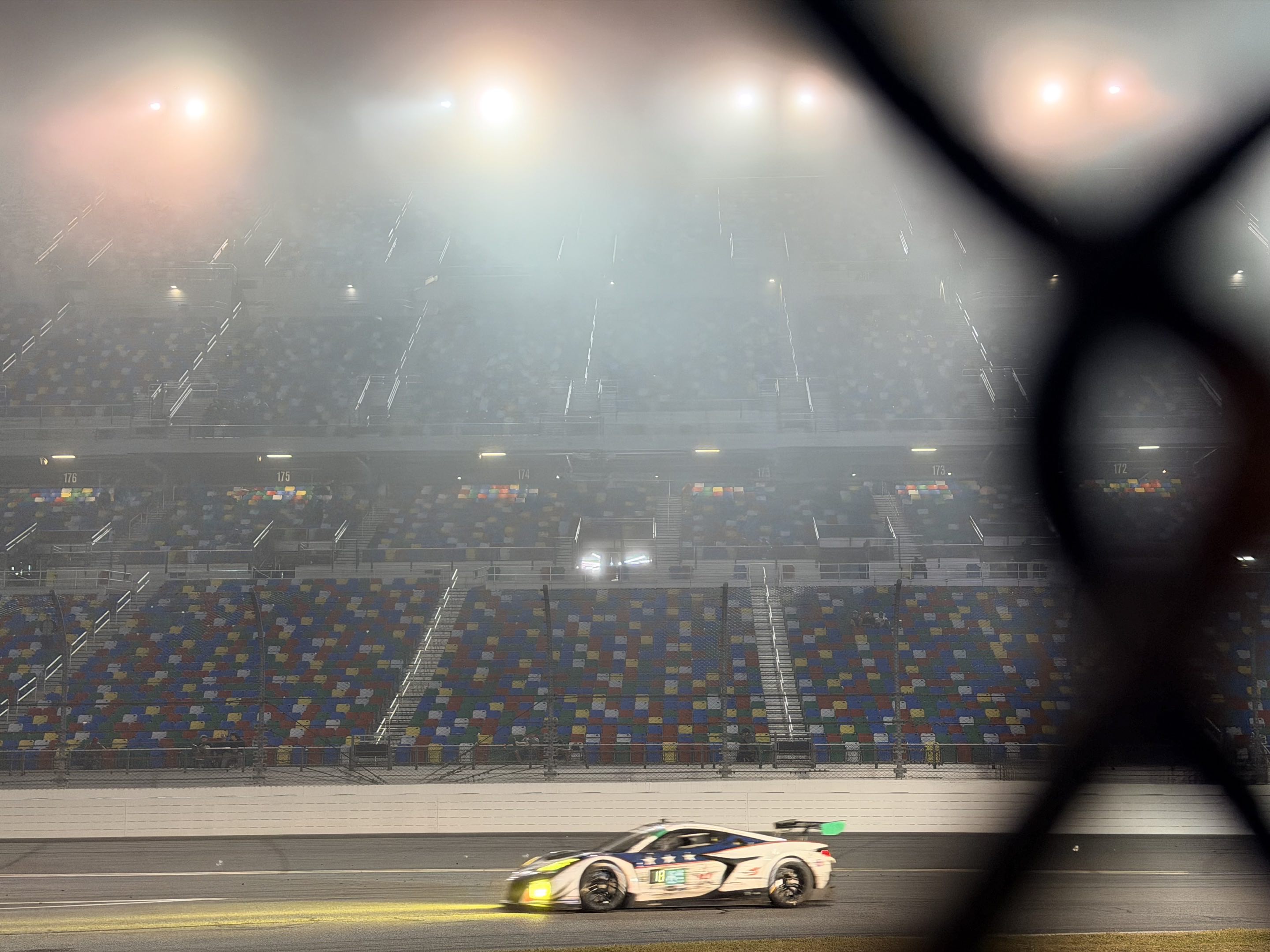 A race car with its headlights illuminating the track in front of it driving past empty grandstands in dark fog backlit by floodlights, viewed through a chain link fence
